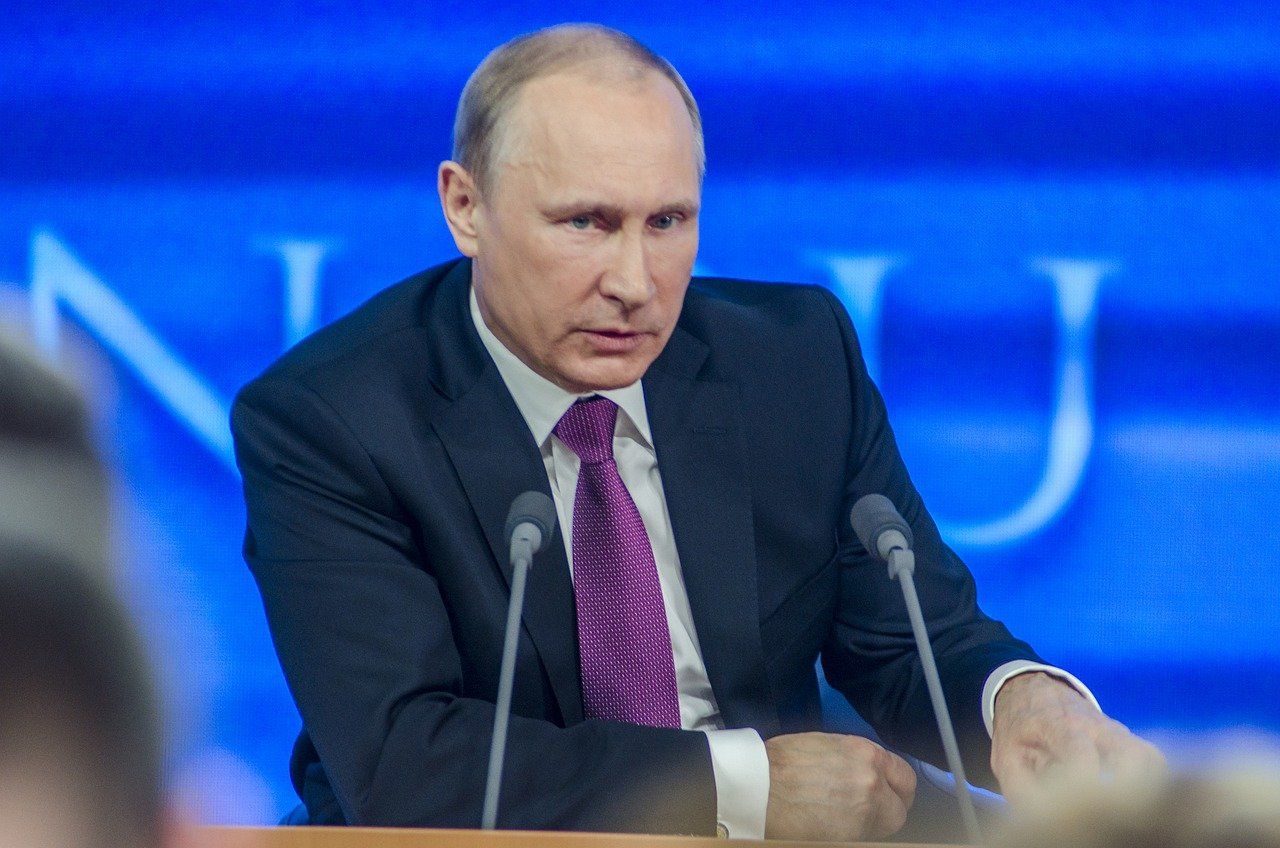 A balding white man in a suit sits behind microphones. In front of him is a sign reading 